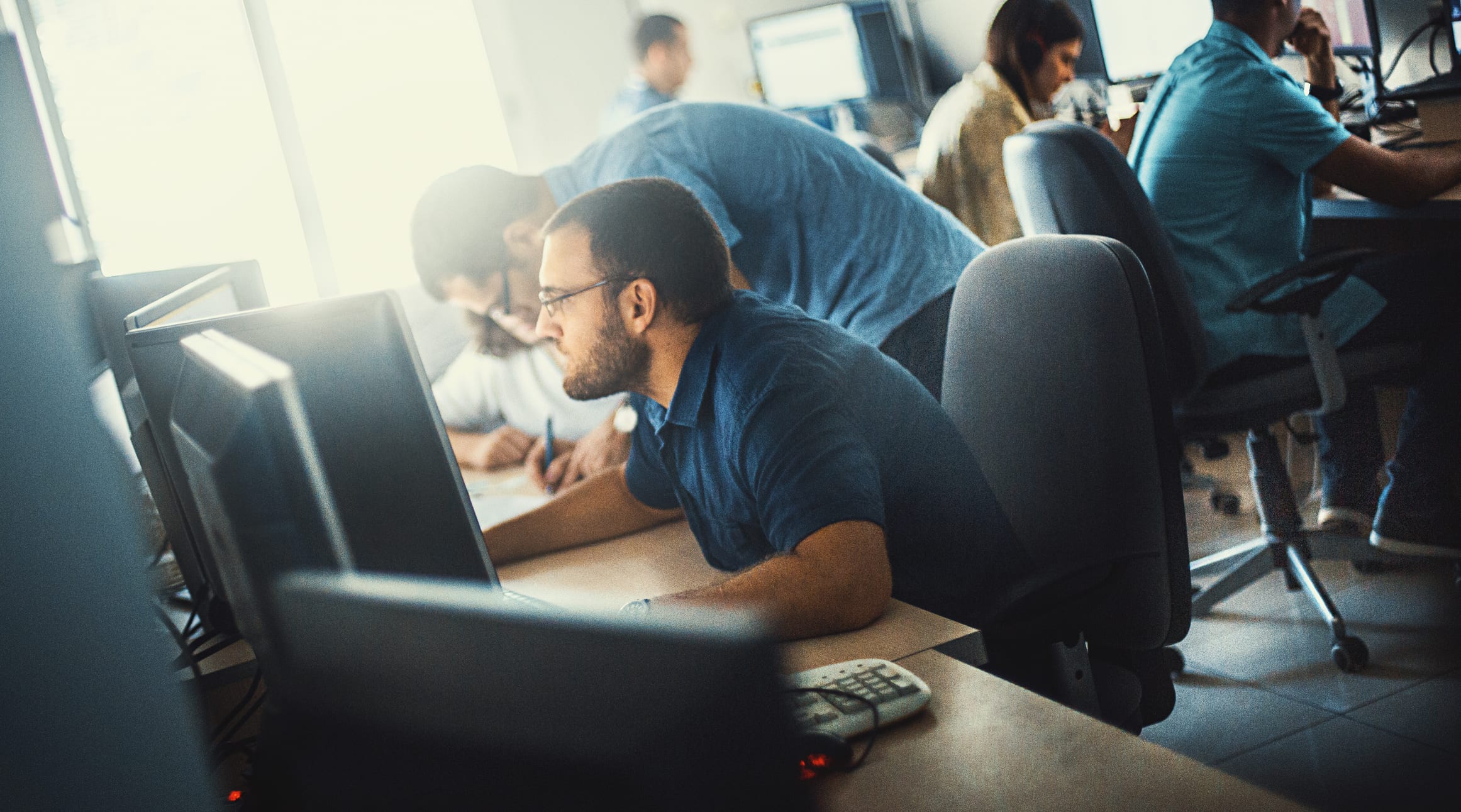 Group of mid 20's and 30's men and women engaged in application development job. They are seated by long desks back to back, each person in front of dual screen computer. 