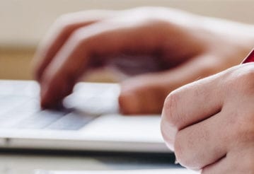 Close up of hands. One is typing, the other holds a pen.