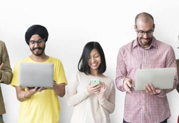 Smiling young adults standing in a line looking at their electronic devices.