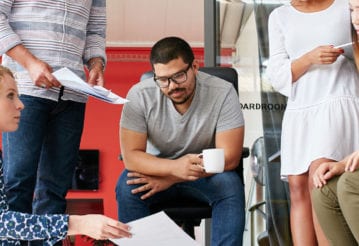 Coworkers sit and stand, looking at a paper held by a woman.