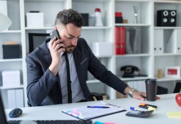 A business man holds a cell phone to his ear while sitting at at desk.