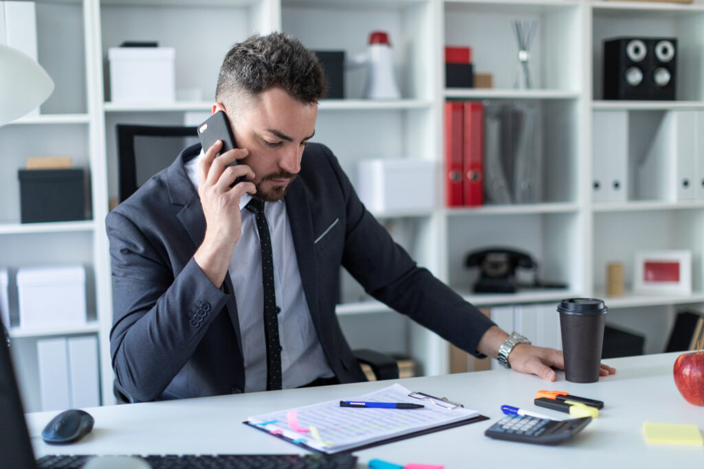 A business man holds a cell phone to his ear while sitting at at desk.
