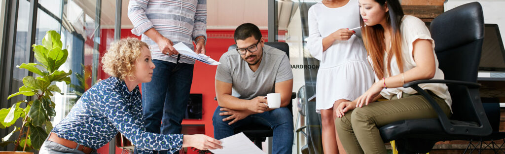 Coworkers sit and stand, looking at a paper held by a woman.