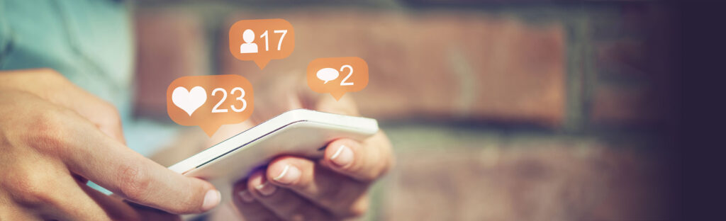 Hands holding a mobile phone that has social media likes, comments, and followers icons floating above it.