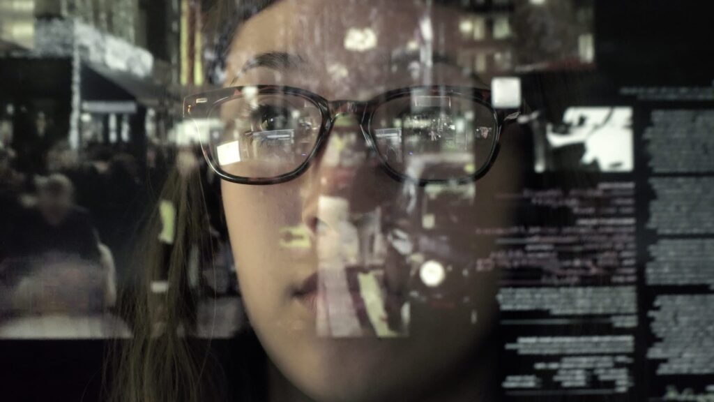 An asian woman concentrating on a touch screen display. The point of view is from behind the screen, looking through the data and images to the woman's face as she manipulates the windows of information.